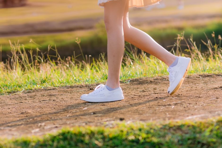 A woman walks on a dirt path in a sunlit park, wearing white sneakers and a skirt.