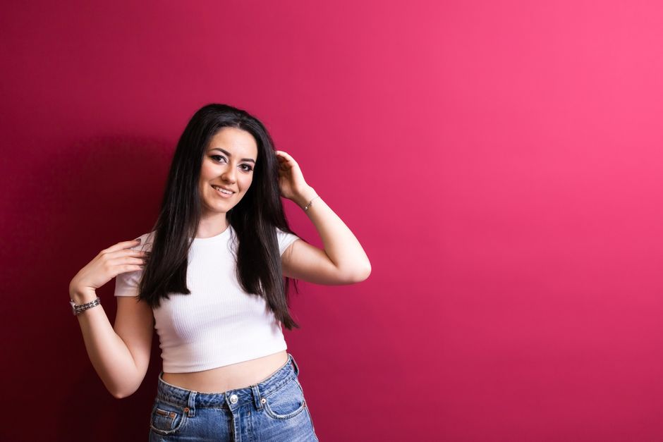 Smiling woman with long black hair wearing a crop top and jeans against a vibrant red background.