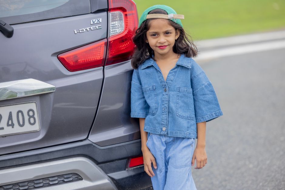 Young girl in denim outfit poses beside a car on a roadside, wearing a green cap.