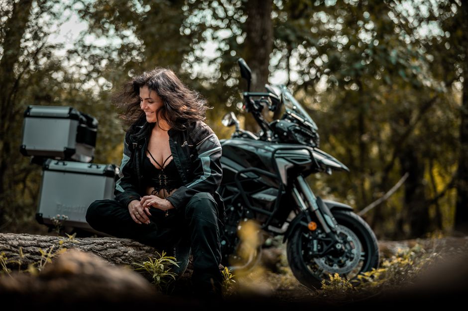 Woman in leather outfit with motorcycle in autumn forest, smiling.