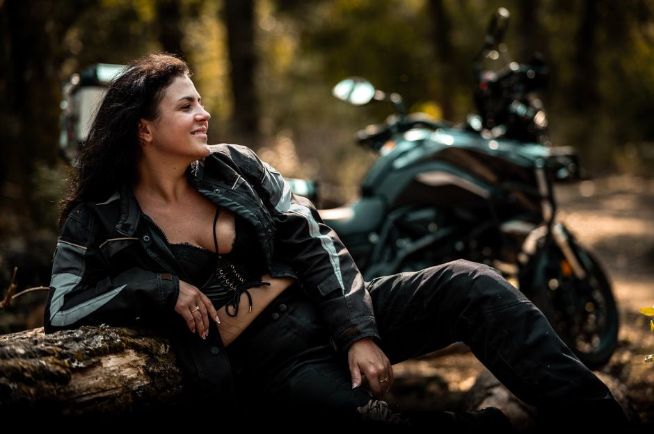 Woman in motorcycle gear relaxing in a forest, smiling near bike.