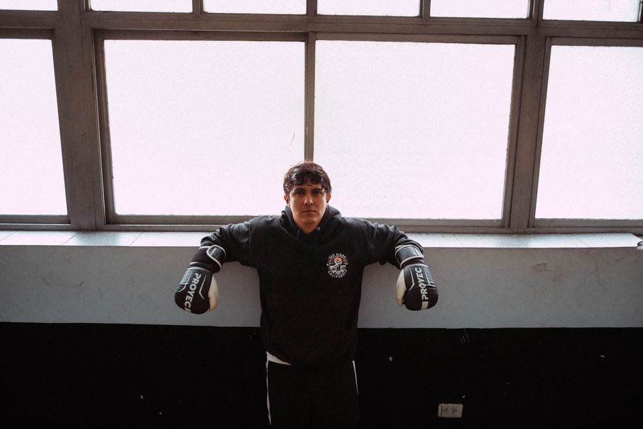 A young boxer stands confidently against a window, ready for training in Buenos Aires gym.