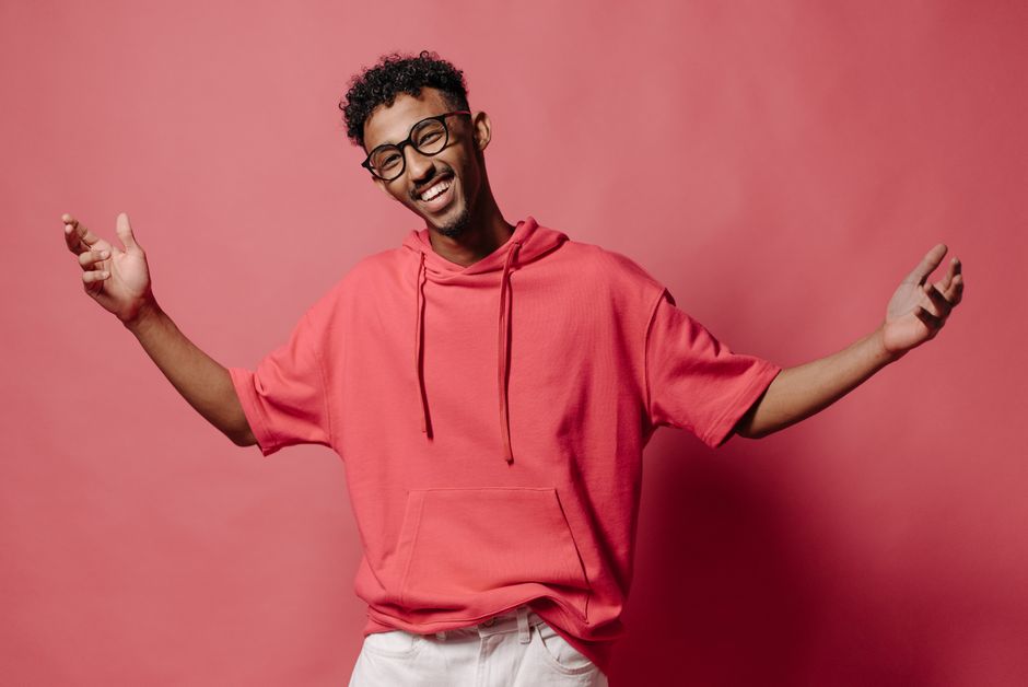 Happy man in a pink hoodie smiles joyfully with arms spread in studio setting.