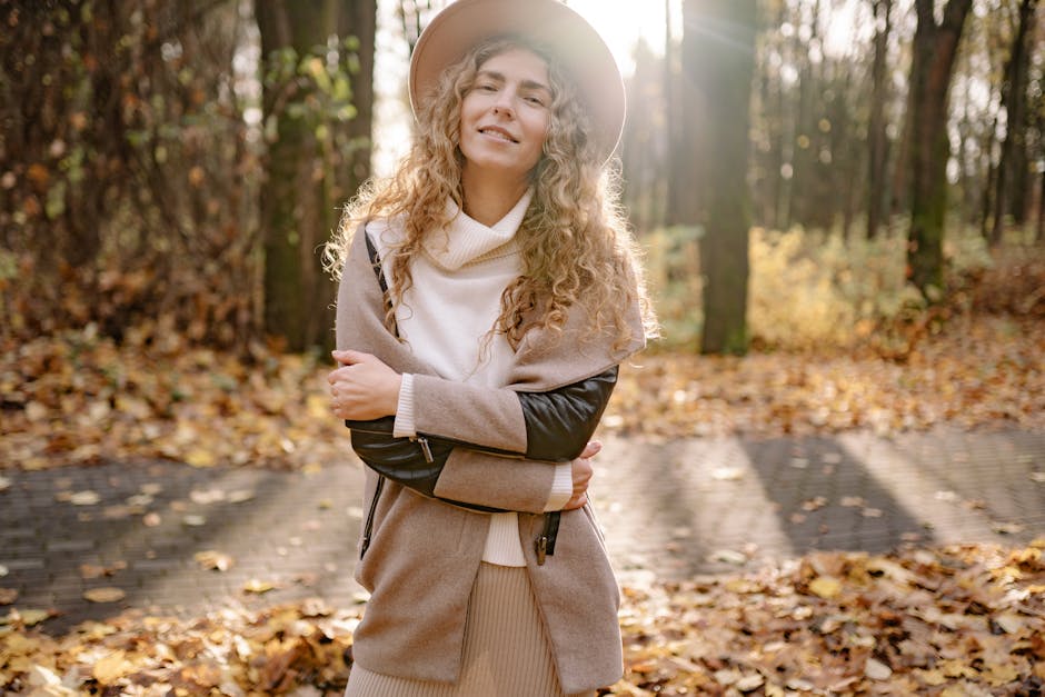 Woman in autumn outfit with hat smiles in a sunlit forest path with fallen leaves.