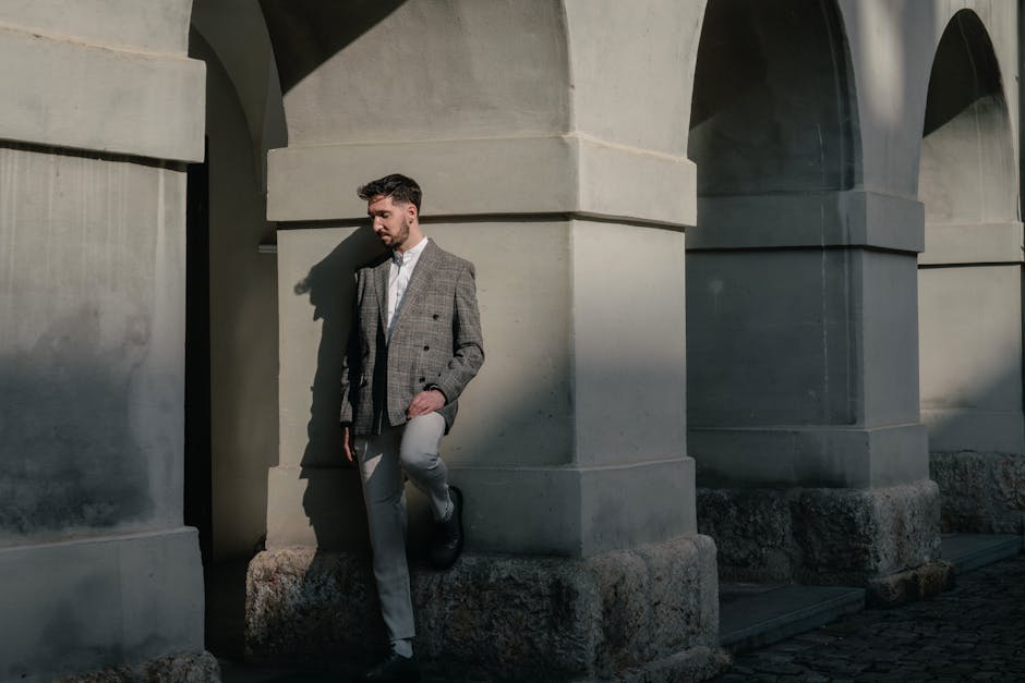 A man in a classic plaid blazer poses by a stone archway in natural light, showcasing modern street fashion.