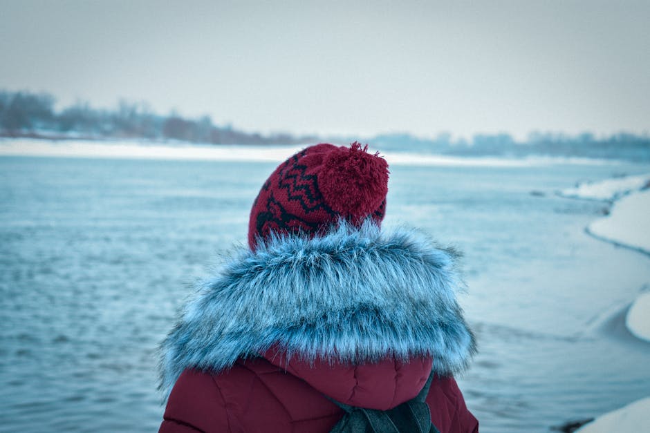 Person in a red beanie and fur coat gazing at a frozen lake during winter.