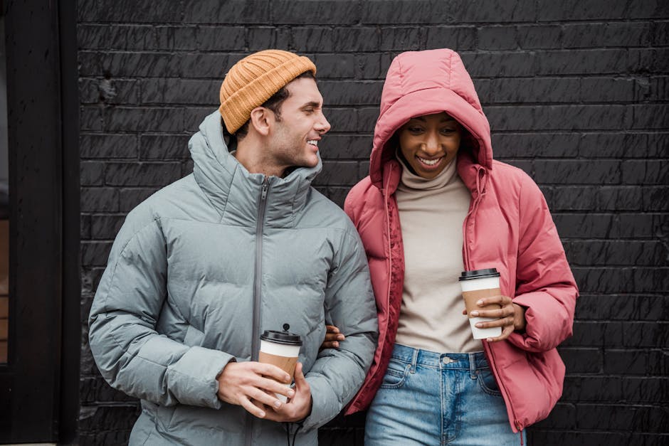 Positive multiethnic couple in outerwear standing close with cups of takeaway coffee on street