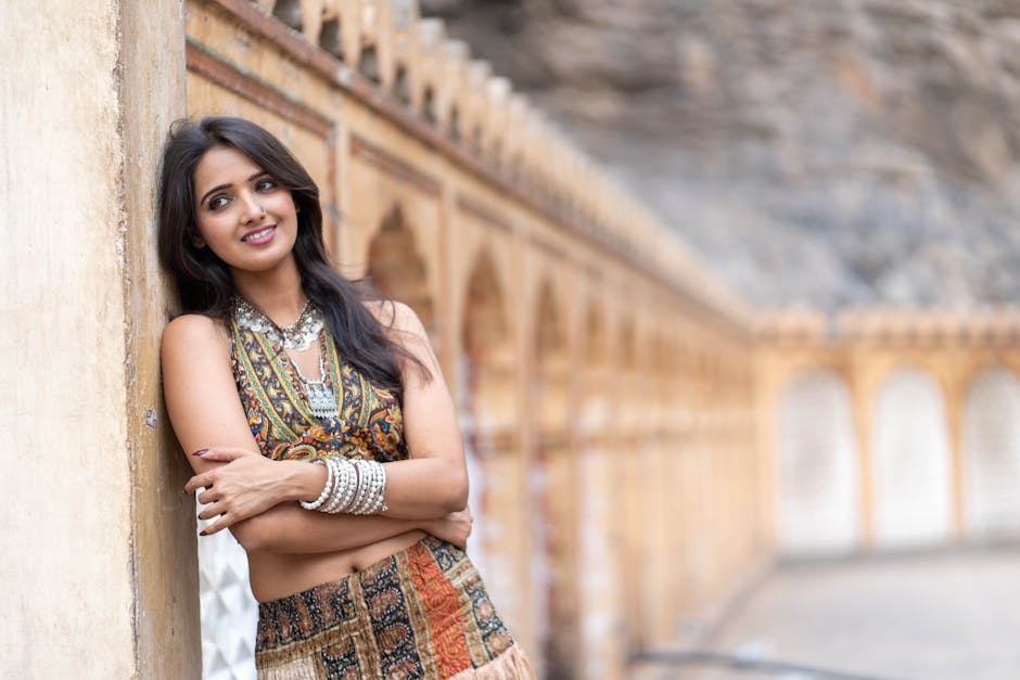A woman in vibrant ethnic attire with silver jewelry poses by a historic wall.