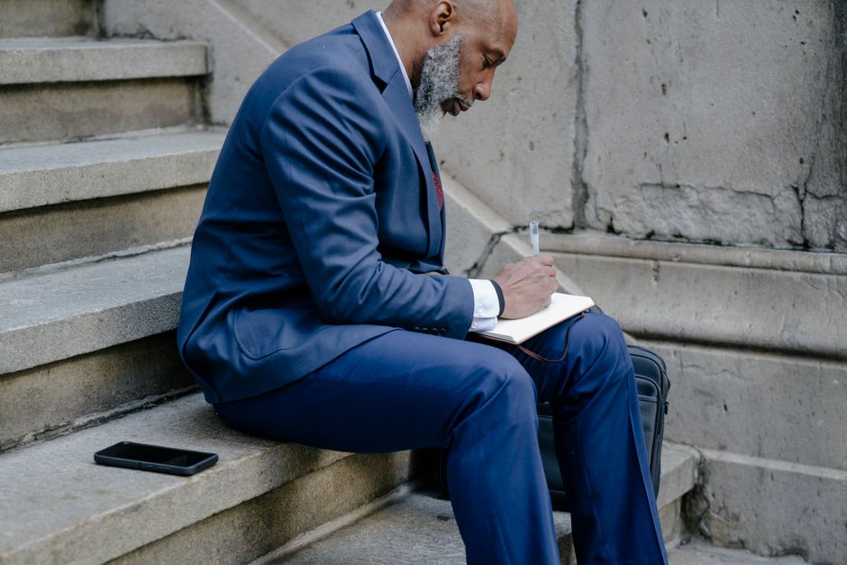 African American man in a suit writing in a notebook while seated on stone stairs.