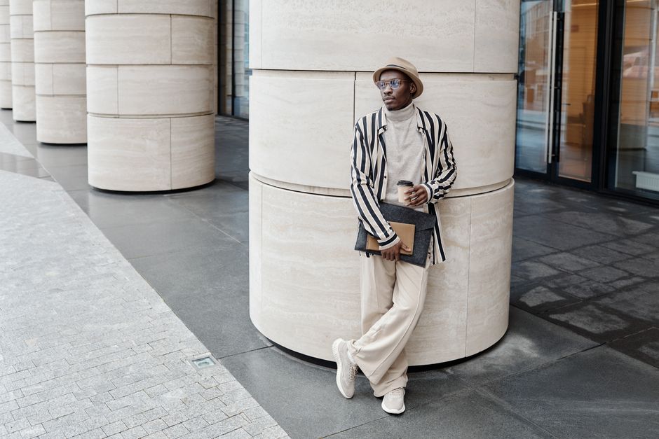 A stylish man in casual attire leans against large modern columns, holding a coffee and folder.