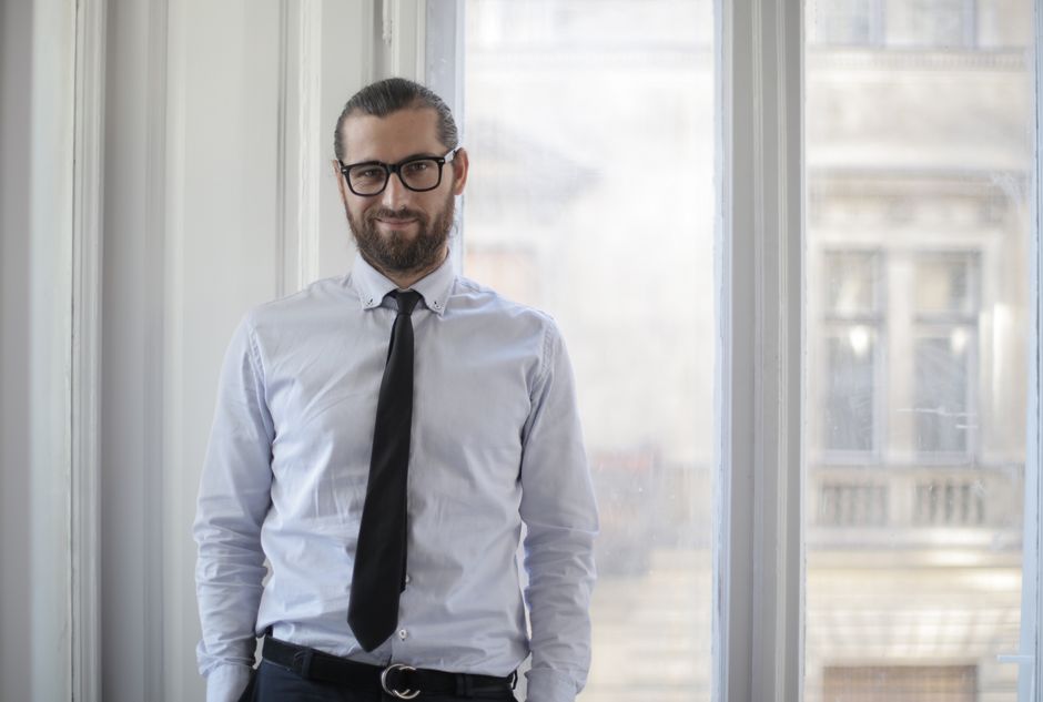 Confident man with glasses and beard posing by a window in a formal setting.