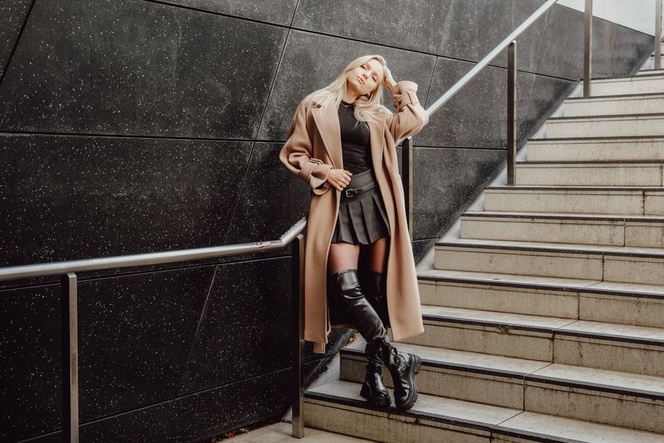 Chic woman poses in a trench coat and knee boots on stylish urban stairs.