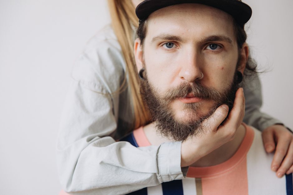 Close-up of a serious bearded man in a cap and striped shirt with a hand touching his face