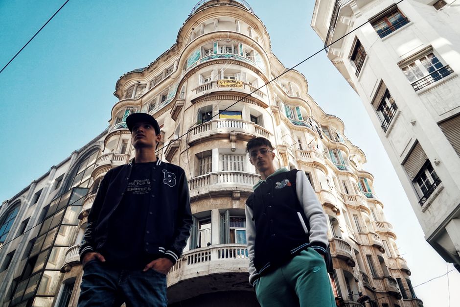 Two young adults in street fashion pose in front of a historic building with ornate balconies.
