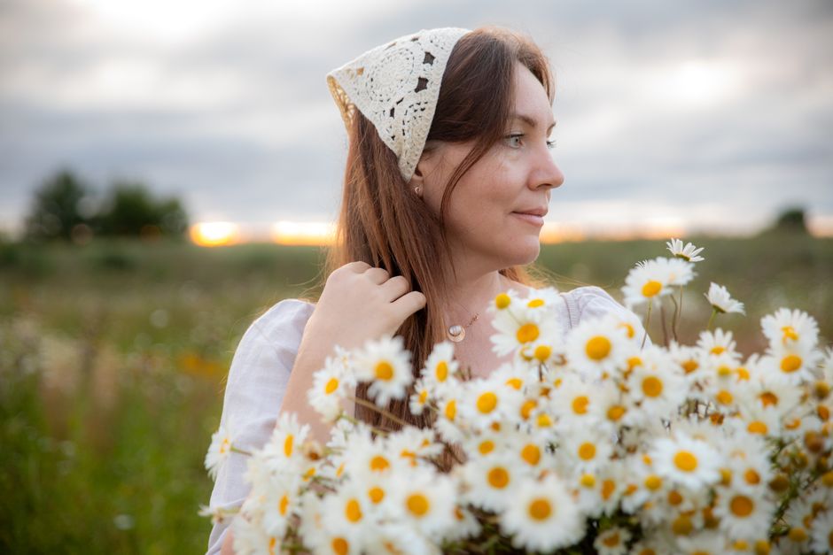 Woman with daisies in a field, wearing a crochet headscarf, during sunset.