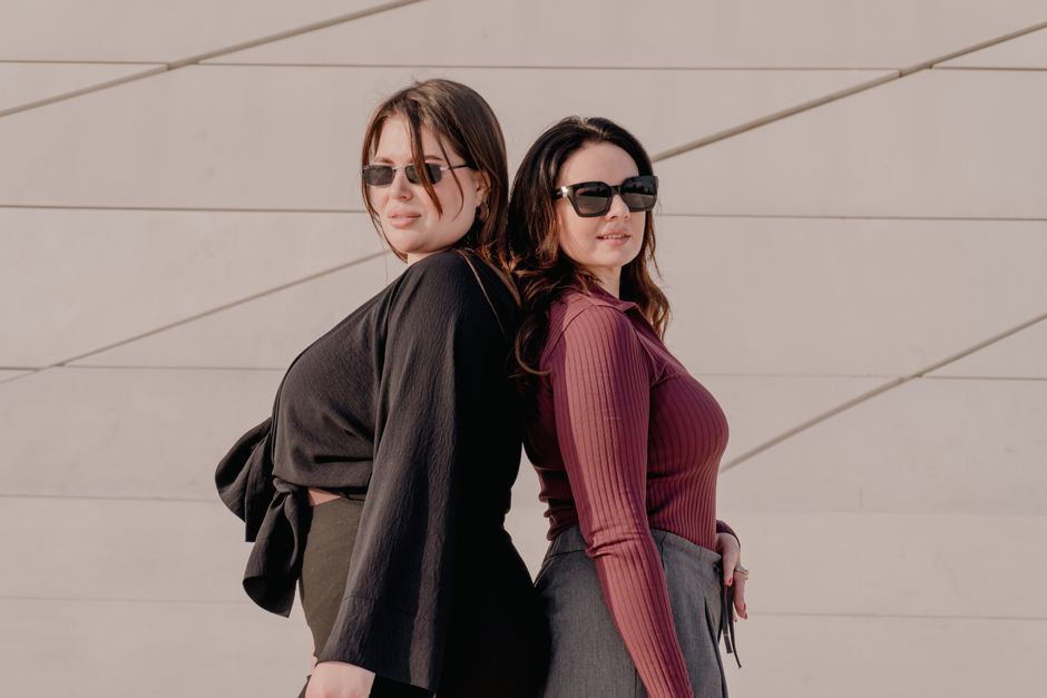 Two stylish women in sunglasses pose back to back against a minimalist wall.