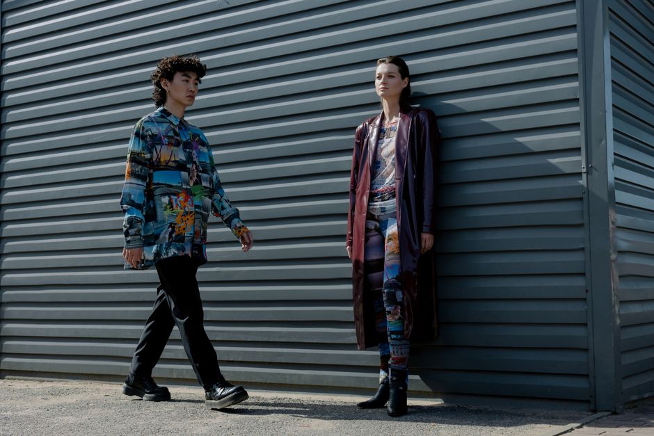 Two models in dynamic streetwear pose in front of an industrial wall.