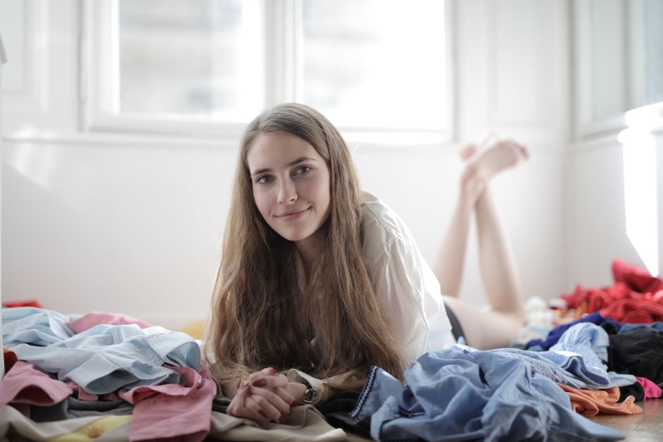 Glad female shopaholic in casual wear lying on floor in untidy room with scattered clothing items while choosing outfit and looking at camera