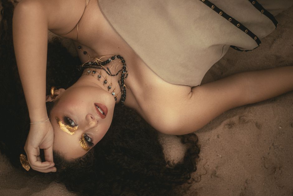 Elegant model posing with gold leaf makeup and necklaces on sandy beach.