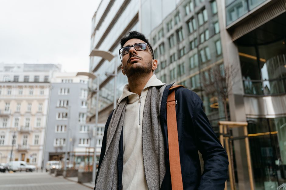 Confident man with scarf and glasses walking through urban city with modern buildings.