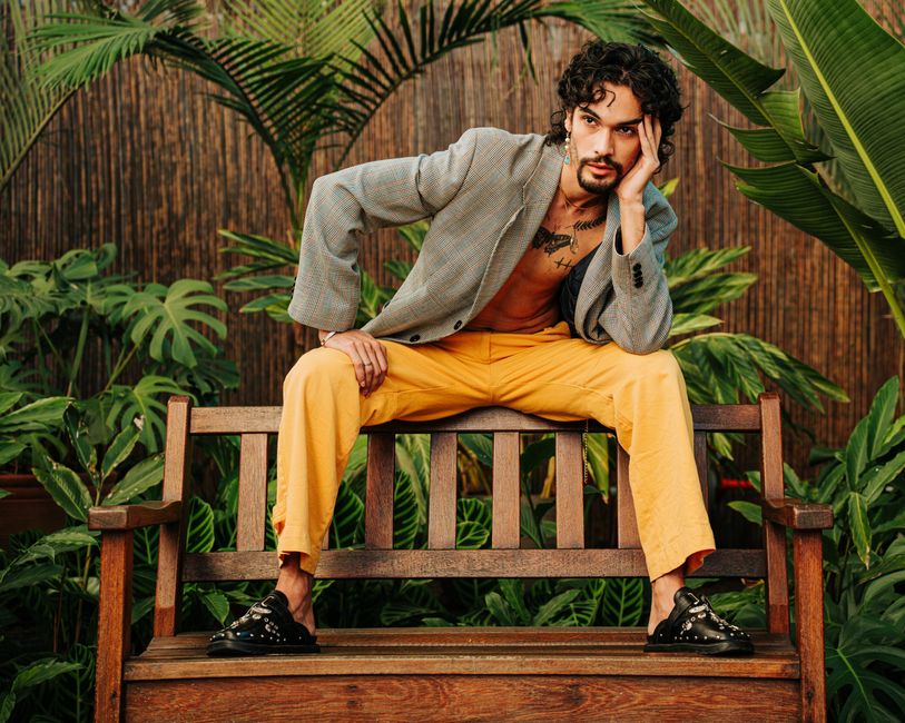 Fashionable man sitting on a wooden bench surrounded by tropical plants, showcasing contemporary style.