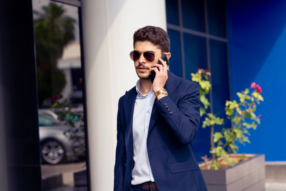 A young professional in sunglasses makes a call outside a modern office building.