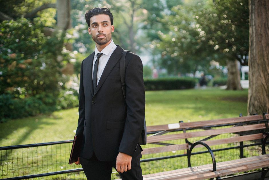 A well-dressed man in a black suit stands confidently in a sunny park.