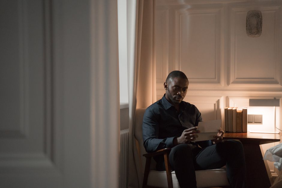A thoughtful man reads a letter while sitting in a cozy room with soft lighting.