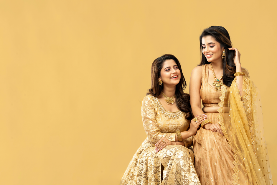 Two smiling women in elegant traditional Indian dresses with a warm backdrop.