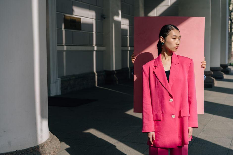 A fashionable woman in a pink blazer stands with a pink backdrop in an architectural setting, using dramatic shadows.