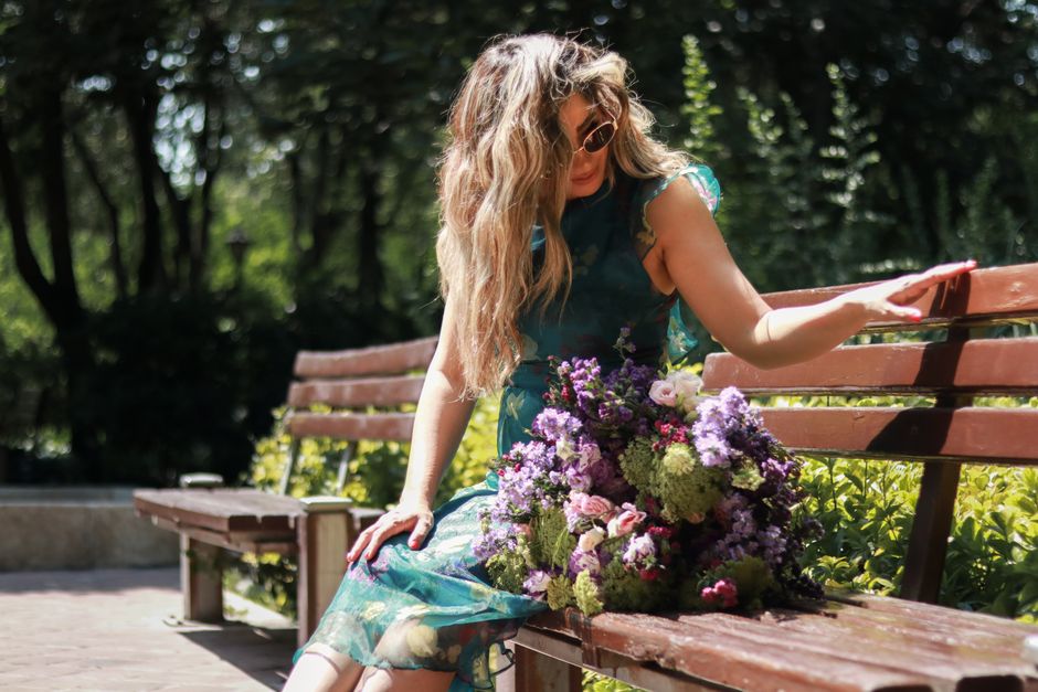 Elegant woman in a floral dress sitting on a park bench with flowers, enjoying a sunny day.