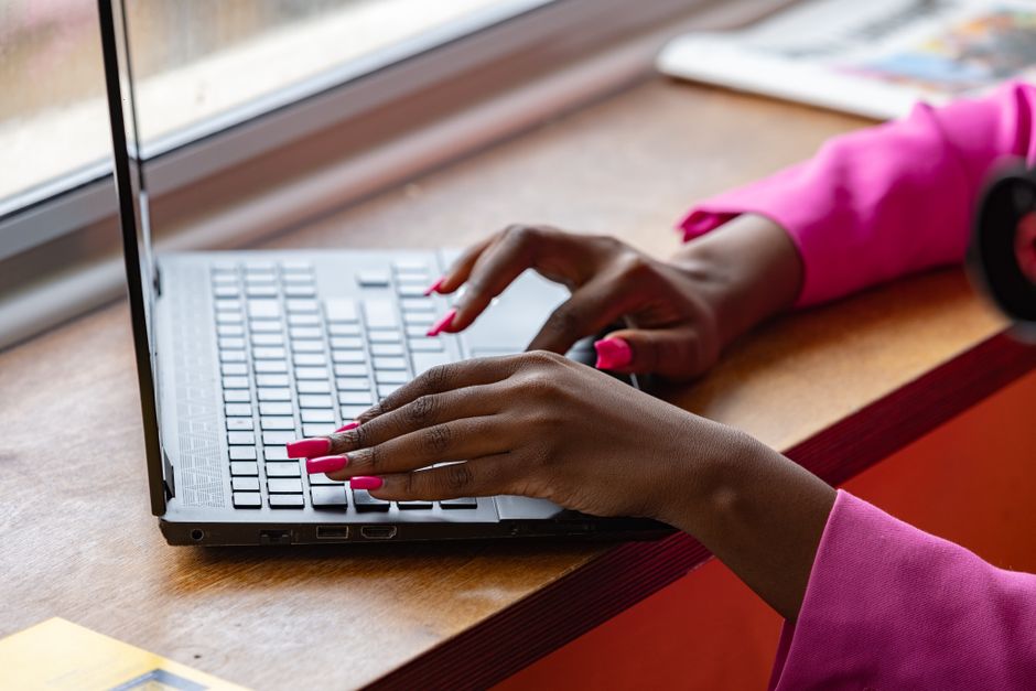 A woman with vibrant pink nails types on a laptop near a window, wearing a bright pink sleeve.