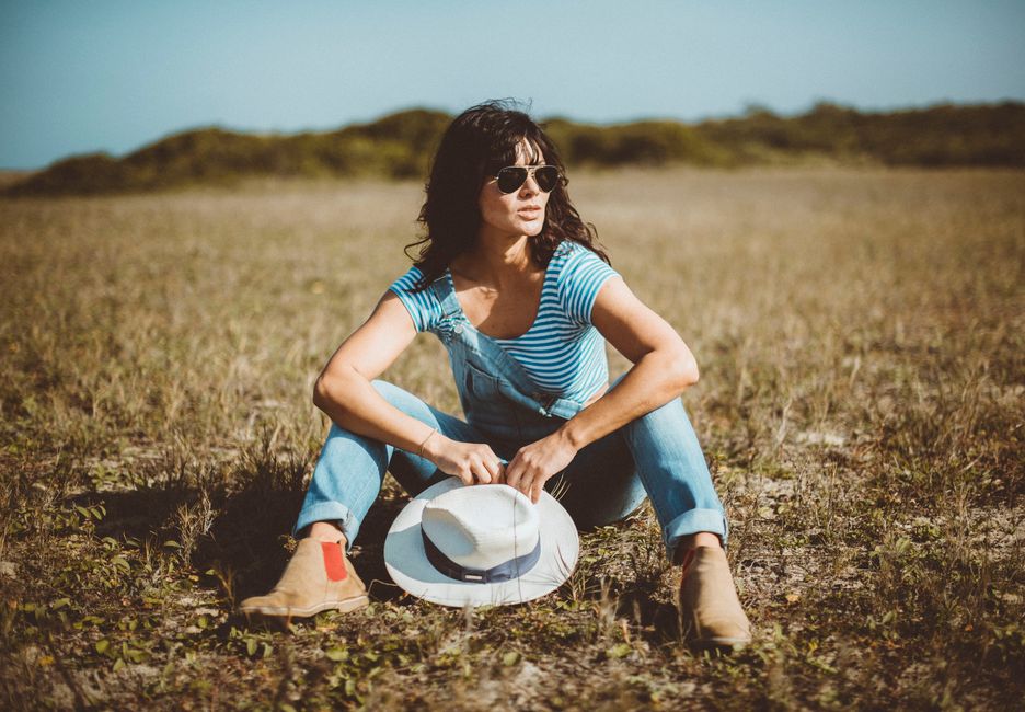 Woman wearing sunglasses and striped top, sitting in a field holding a hat. Fashion photography in rural setting.