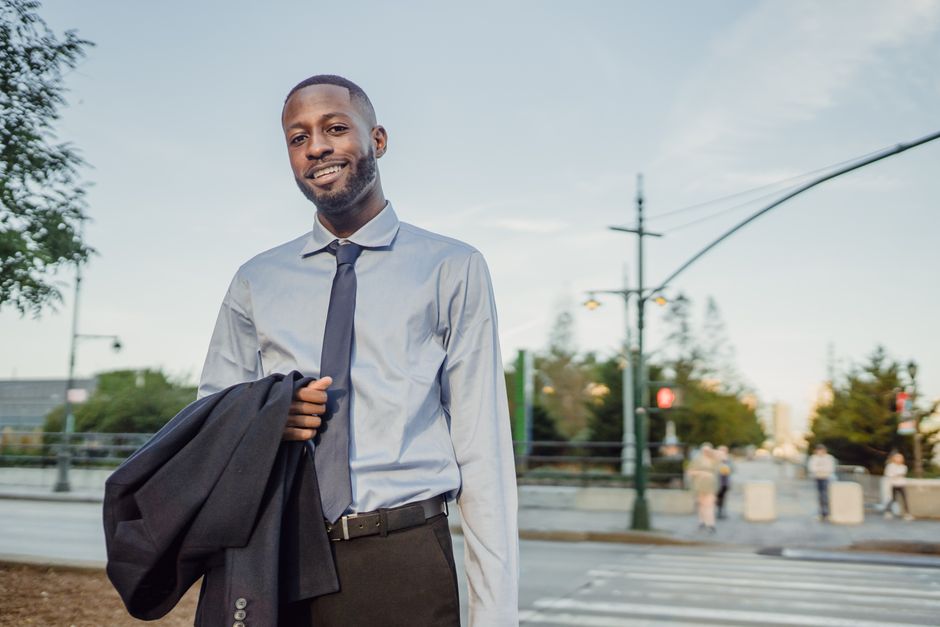 Businessman in formal attire smiling and holding a suit jacket outdoors in an urban setting.
