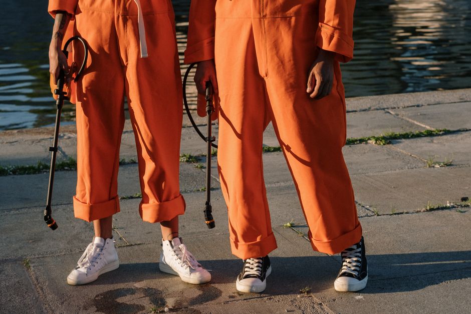 Two individuals in orange jumpsuits holding sprayers on a concrete surface by the water.
