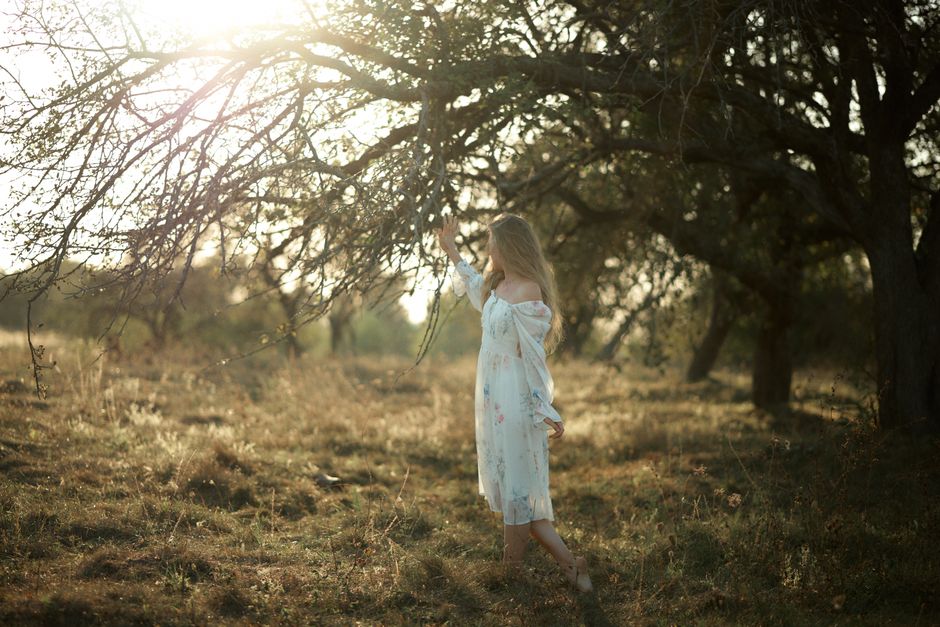 Woman in white dress enjoying sunlight in meadow under a tree.