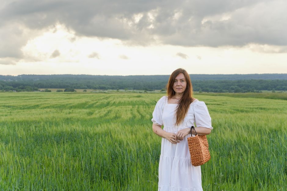 A woman wearing a white dress with a basket stands in a lush green field under a cloudy sky.