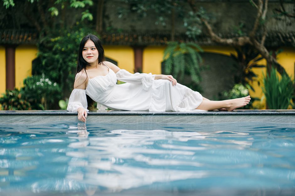 Full body of positive barefoot Asian female in white dress looking away while lying on edge of swimming pool near building