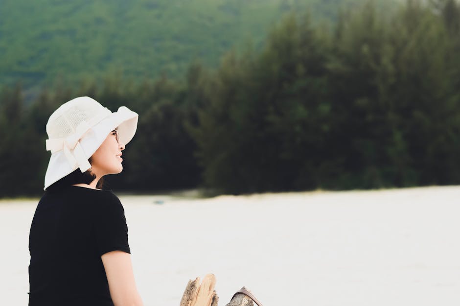 A woman in a white sun hat enjoying a serene moment on a sandy beach with lush greenery.