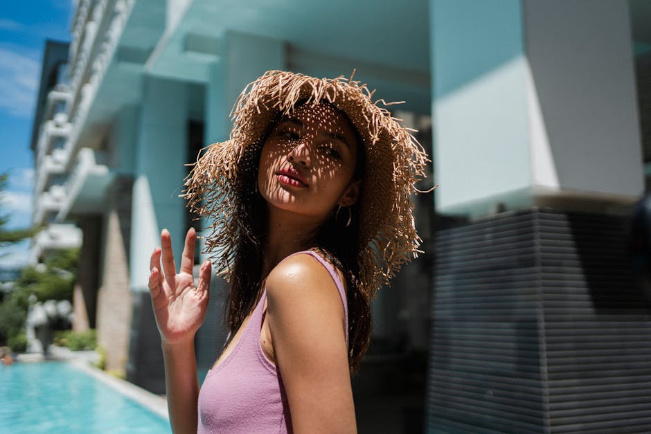 Side view of stylish young Asian female in light top and straw hat chilling near swimming pool in yard of modern hotel on sunny day
