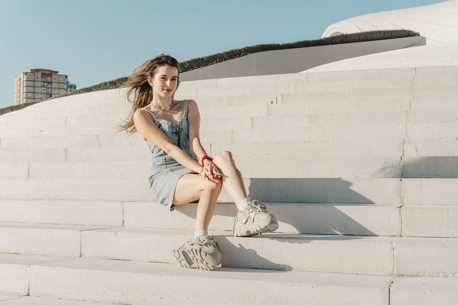 Young woman in casual attire sitting on contemporary white steps under blue sky, capturing a relaxed outdoor moment.