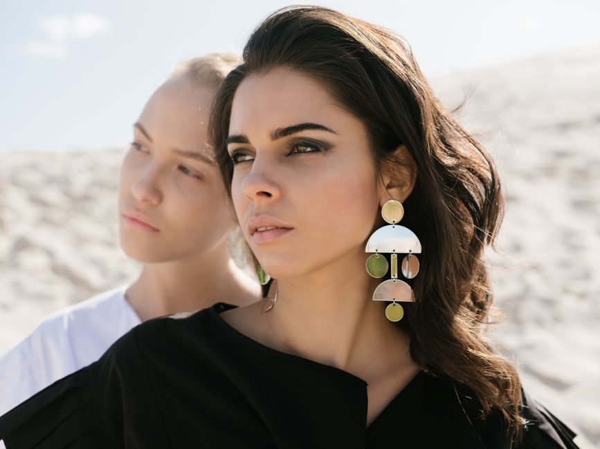 Serene brunette woman with elegant wavy hairstyle wearing black dress with fashionable earrings and makeup standing in front of young blond female friend with simple hairstyle and without makeup while looking away