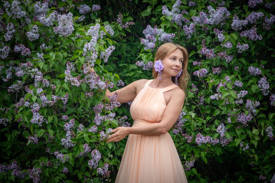 A woman in a peach dress stands gracefully among lilac bushes, radiating elegance and natural beauty.