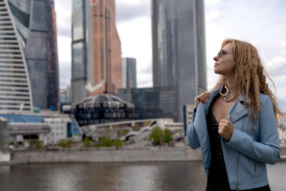 A confident woman in a blue jacket poses against a modern urban cityscape with skyscrapers.