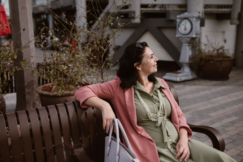 Woman in pink blazer and green dress sitting on a bench outdoors, gazing thoughtfully.