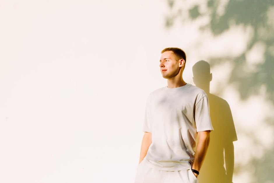 Young man standing against a sunlit wall with hands in pockets, casting a shadow.