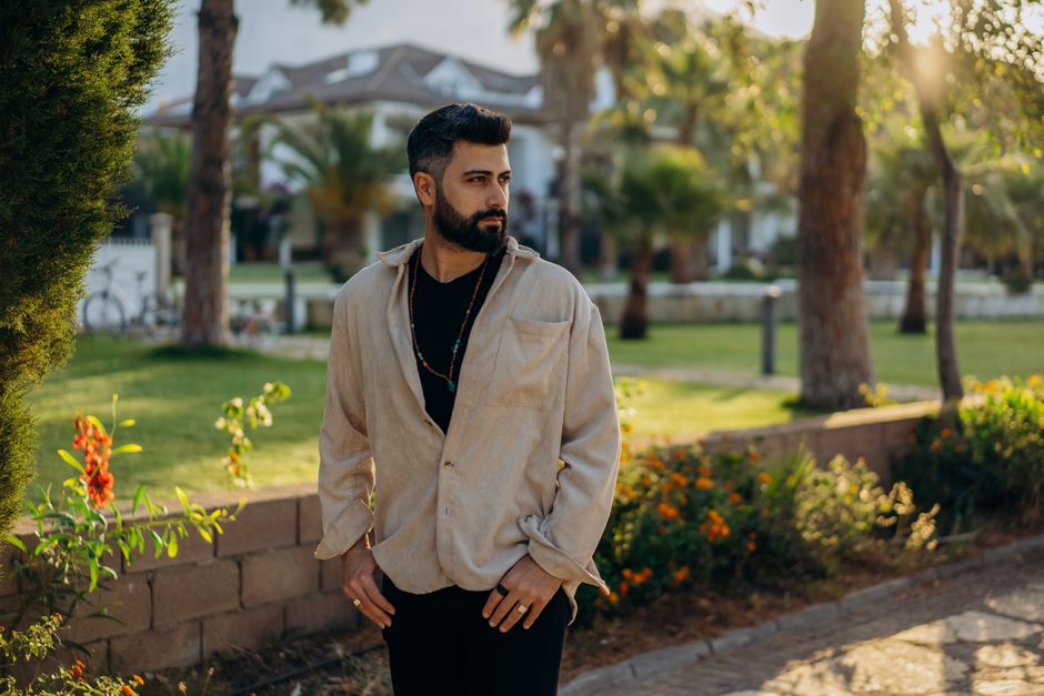 A fashionable man with facial hair standing in a sunlit park setting.