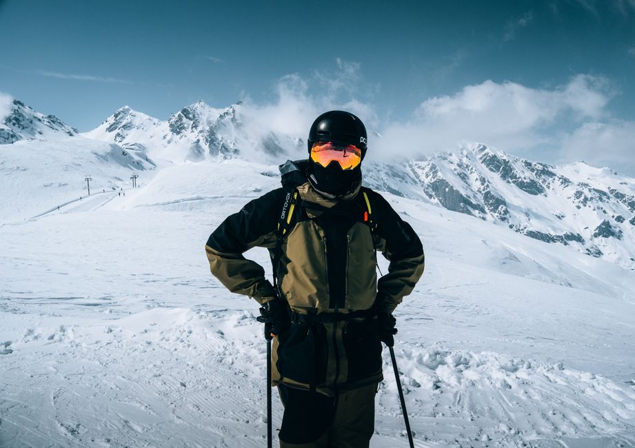 A skier stands confidently in full gear against a backdrop of snow-covered alpine peaks.