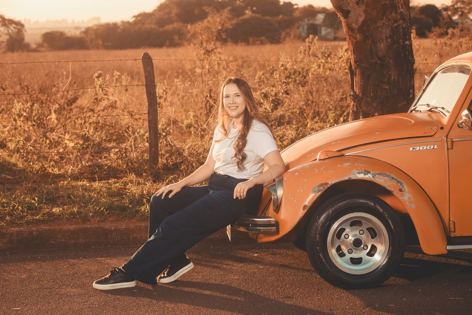 A woman enjoys a sunny day sitting beside a classic orange car in a rural setting.