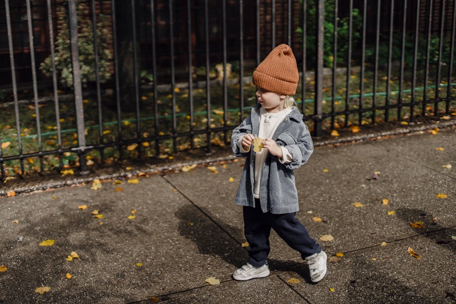 From above full body side view of cute kid in hat standing on walkway near metal fence with dried leaf in hand on autumn day